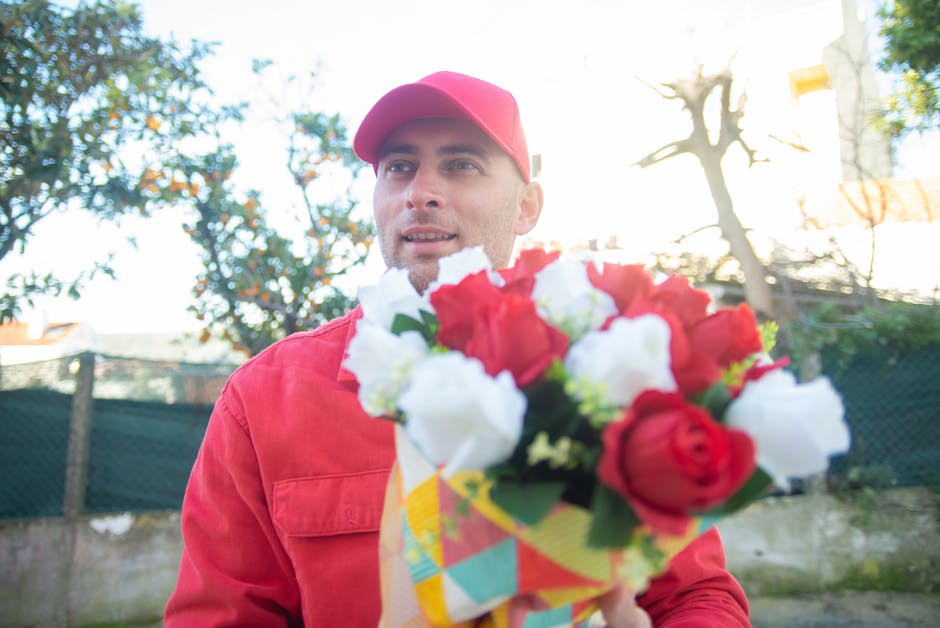 A male florist wearing a red cap and matching red jacket holds a vibrant bouquet of fresh flowers, featuring red and white roses with green foliage, wrapped in colorful paper. He is standing outdoors
