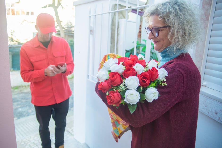 A woman with curly gray hair and glasses is holding a vibrant bouquet of fresh red and white roses, arranged with green foliage, wrapped in colorful patterned paper. She stands near a door or window,