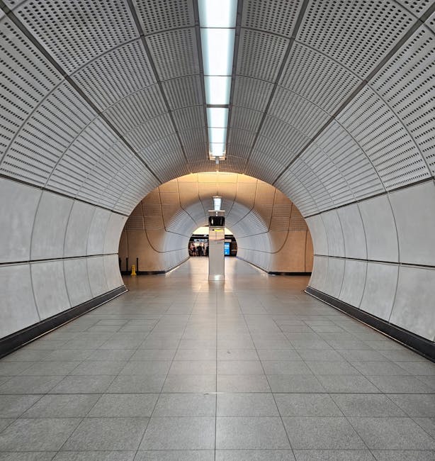 The image shows a modern underground tunnel at South Tottenham station with a curved, metallic ceiling featuring a perforated pattern, illuminated by a linear light fixture running along its center. T