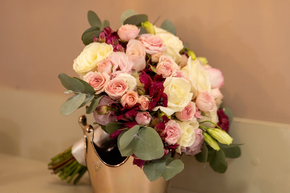 A bouquet of fresh flowers arranged in a compact, rounded style sits in a reflective silver vase. The bouquet contains pastel pink roses, ivory roses, and cream-colored lisianthus, accented with deep 